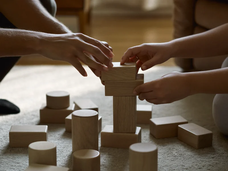 Erwachsenen- und Kinderhände beim gemeinsamen Bauen mit Holzklötzen am Boden